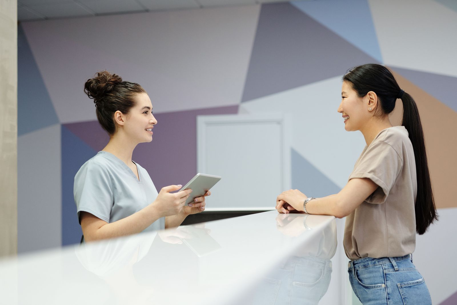 A front-desk coordinator with a tablet speaks with a patient at a clinic reception counter.